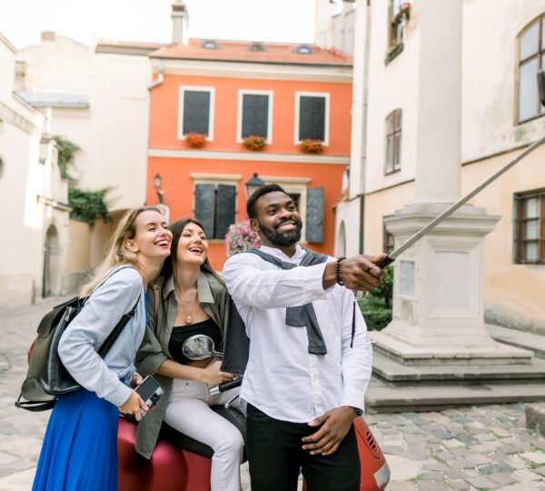 Multiethnical happy young people, African guy and two Caucasian girls, making selfie photo with funny laughing faces, standing in the old city yard with beautiful ancient buildings.